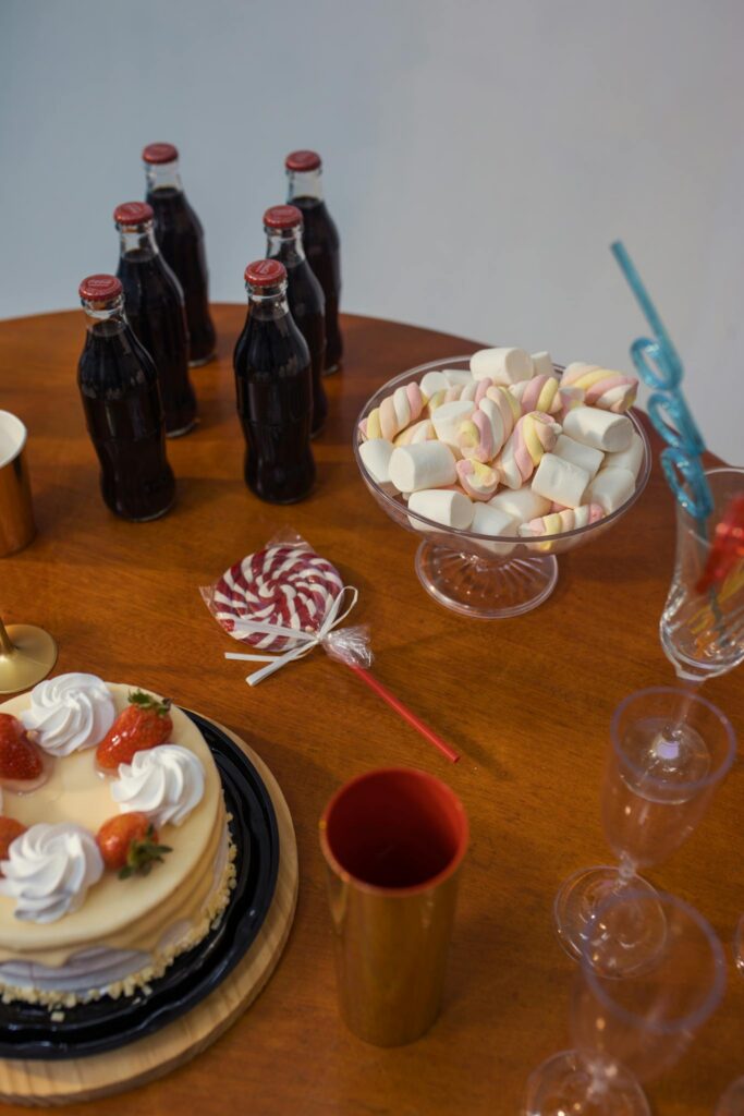 Capture of a Brazilian birthday setup with cake, soda bottles, marshmallows, and party items on a wooden table.